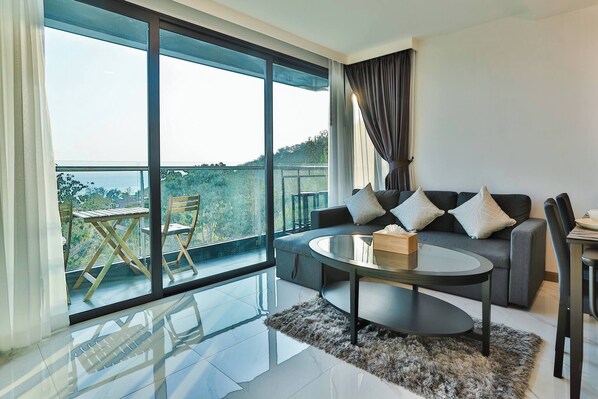 A modern living room with a glass floor-to-ceiling window, a grey sofa with white cushions, a round coffee table, and a small wooden table outside.
