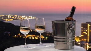 Uma mesa com dois copos de vinho branco e uma garrafa em um recipiente de metal, com uma paisagem urbana ao fundo durante o pôr do sol.