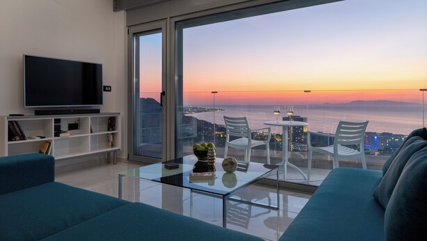 Sala de estar moderna com uma televisão de tela plana grande, uma mesa de centro de vidro e um sofá. Há uma varanda com mesa e cadeiras, oferecendo vista para a cidade e um pôr do sol.