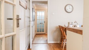 A hallway with a wooden floor leading to a door with frosted glass panels. A wooden chair is placed near a counter with a clock on the wall. - Sea Beach Cottage (Eastbourne)