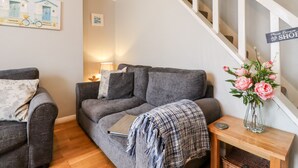 A living room with a grey sofa, a wooden side table with a vase of flowers, and a staircase with a 'Please Remove Shoes' sign. - Sea Beach Cottage (Eastbourne)