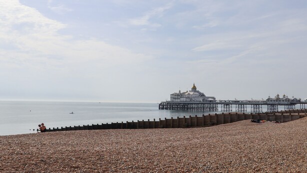 A pebbled beach with a pier extending into the sea, a large building with a golden dome, and a person sitting on the shore. - Sea Beach Cottage (Eastbourne)