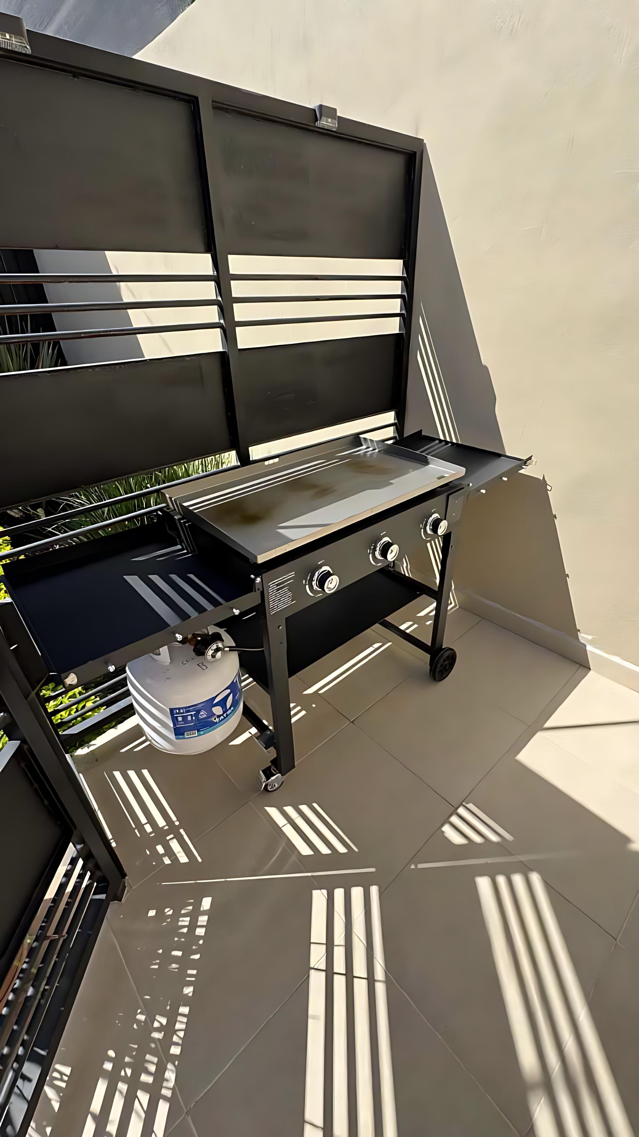 Un barbecue doté d’une structure noire et d’une surface de cuisson en acier inoxydable, placé sur une terrasse carrelée avec un mur blanc en arrière-plan.