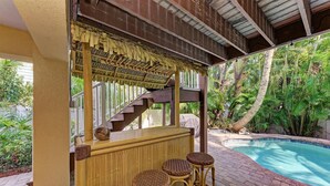 A poolside bar area with wicker stools and a wooden bar, surrounded by lush greenery and a clear pool.