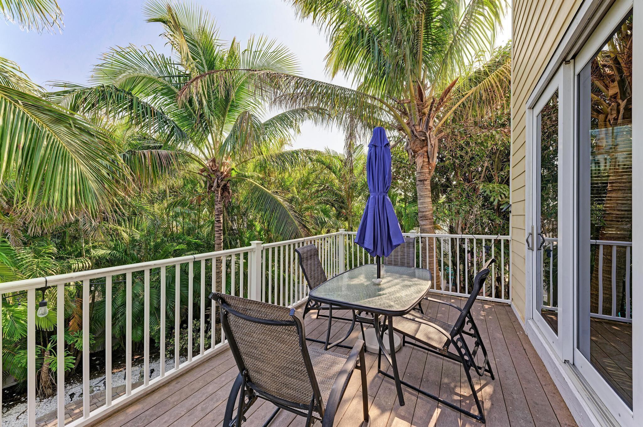 A wooden deck with a glass table and chairs, surrounded by a white railing and palm trees.