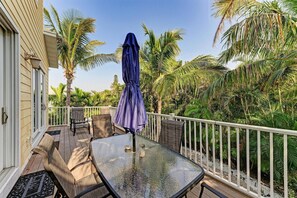 Une table à plateau en verre, avec des chaises et un parasol, sur un balcon doté d’une balustrade blanche, le tout entouré de palmiers.