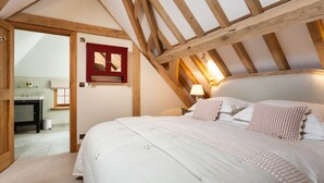 Una camera da letto con soffitto spiovente, travi in legno, un letto con biancheria bianca, un comodino con una lampada e un bagno visibile attraverso una porta aperta.