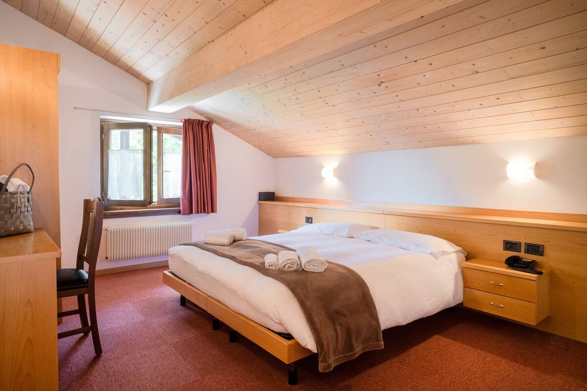 A bedroom with a wooden ceiling, a bed with a brown blanket, a nightstand, a chair, and a window with curtains.