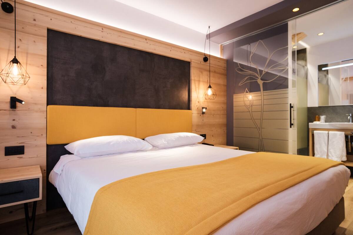 A modern hotel room with a large bed, wooden panel walls, a dark accent wall, and a bathroom with a mirror and towel rack.