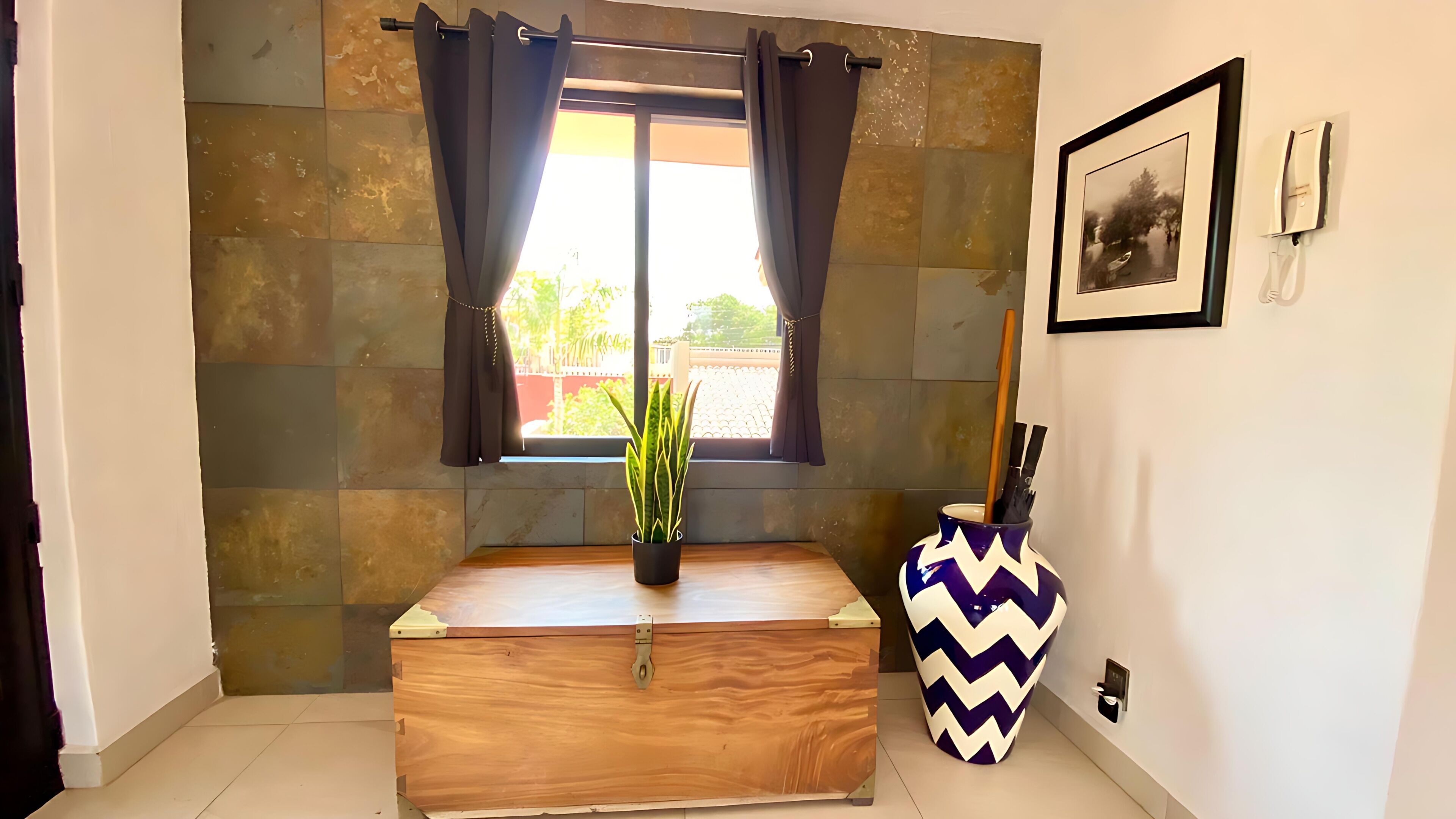 A room with a wooden chest, a potted plant, a decorative vase, and a window with curtains.