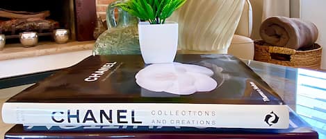Ett vardagsrum med en tv-apparat som visar en film, en öppen spis, en lampa, en krukväxt och en hög med böcker, inklusive titlarna ”Chanel” och ”Louis Vuitton”.
