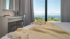 A modern bedroom with a large window, a desk with a hat and vase, and a bed with a textured blanket. - Bianca Luxury Villa - Private Pool (Zakynthos)