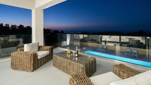A modern outdoor lounge area with wicker furniture, a glass balustrade, and a view of the landscape. - Bianca Luxury Villa - Private Pool (Zakynthos)