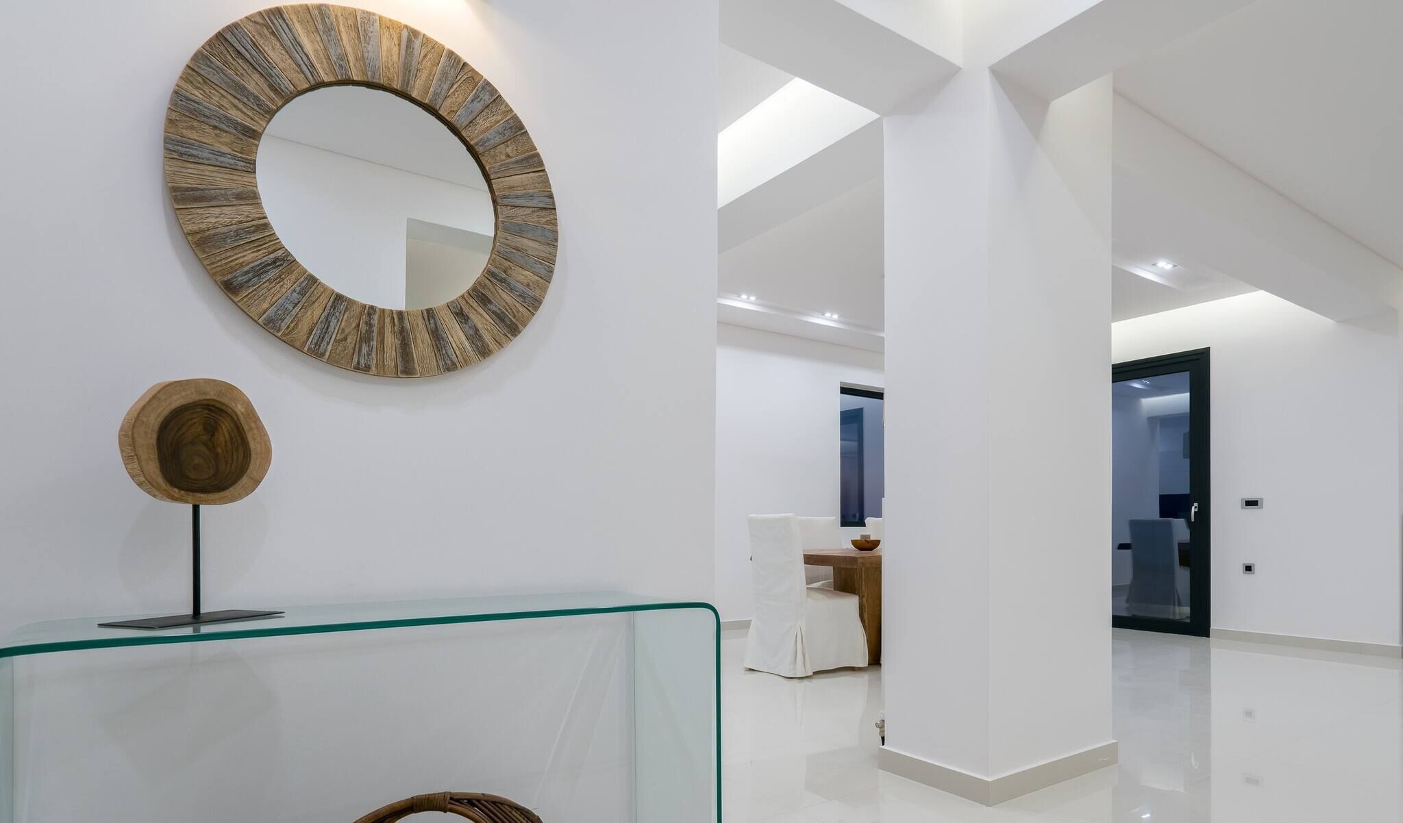 Een modern interieur met een glazen consoles en een houten decoratieve spiegel, en een gang die leidt naar een kamer met een bed en een badkamer.
