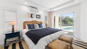 Ein modernes Schlafzimmer mit einem großen Bett, einem Nachttisch, einer Lampe und Blick auf einen Swimmingpool.