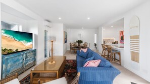 Ein modernes Wohnzimmer mit großem Fernseher, einem blauen Sofa, einem Holztisch und einer Essecke mit Korbstühlen.