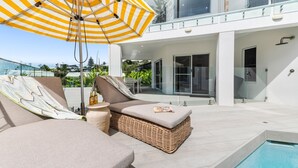 Ein moderner Außenbereich mit Lounge-Möbeln und Pool, mit einer Korbsessel-Couch und einem gestreiften Sonnenschirm.
