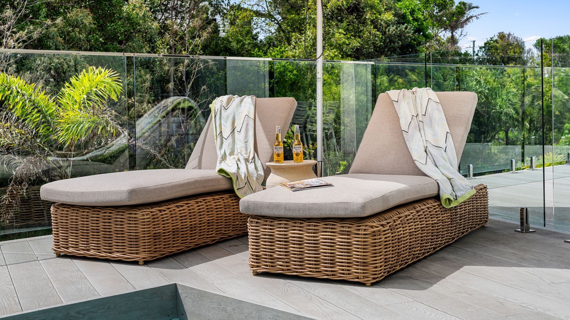 Two wicker lounge chairs with cushions, a small table with bottles, and a glass railing overlooking greenery.