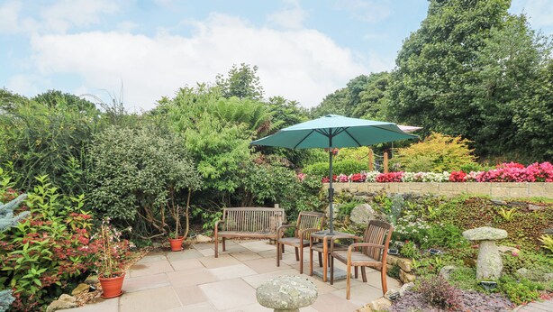 An outdoor patio with a wooden table and chairs, a green umbrella, and various potted plants and flowers. - The Hideaway (St Austell)