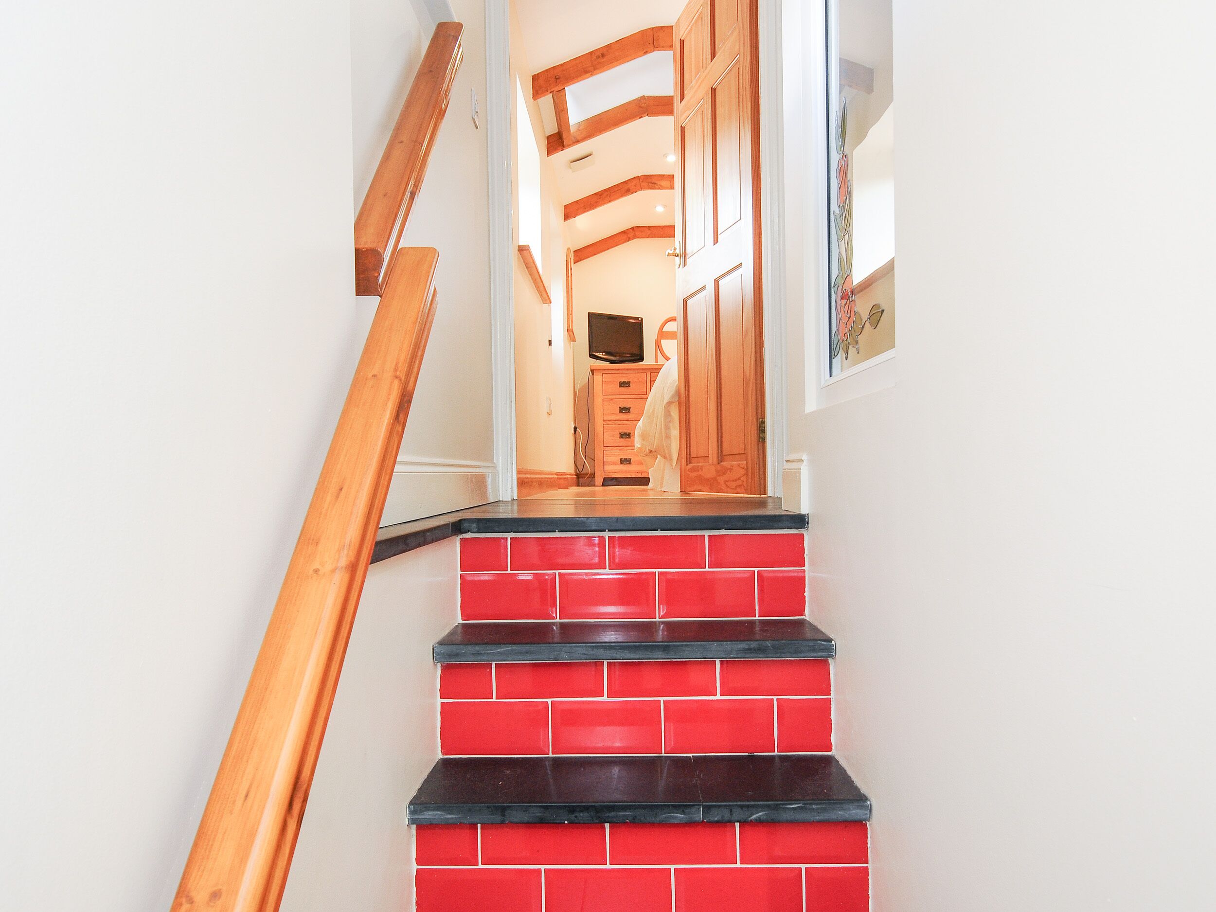 Un escalier aux marches rouges et noires menant à une pièce dotée de poutres en bois et d’un téléviseur.