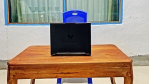 A laptop on a wooden table with blue legs, a blue chair, and a window with curtains in the background. - OYO 91927 Homestay Kebun (Kupang)