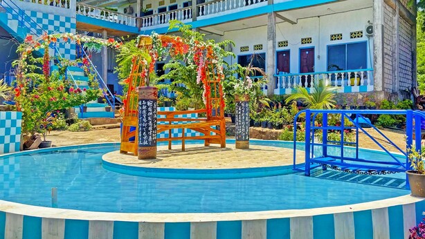 A swimming pool with a wooden deck and a blue and white building in the background. - OYO 91927 Homestay Kebun (Kupang)