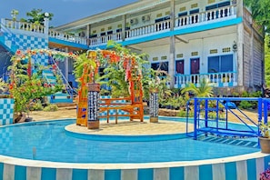 A swimming pool with a wooden deck and a blue and white building in the background. - OYO 91927 Homestay Kebun (Kupang)