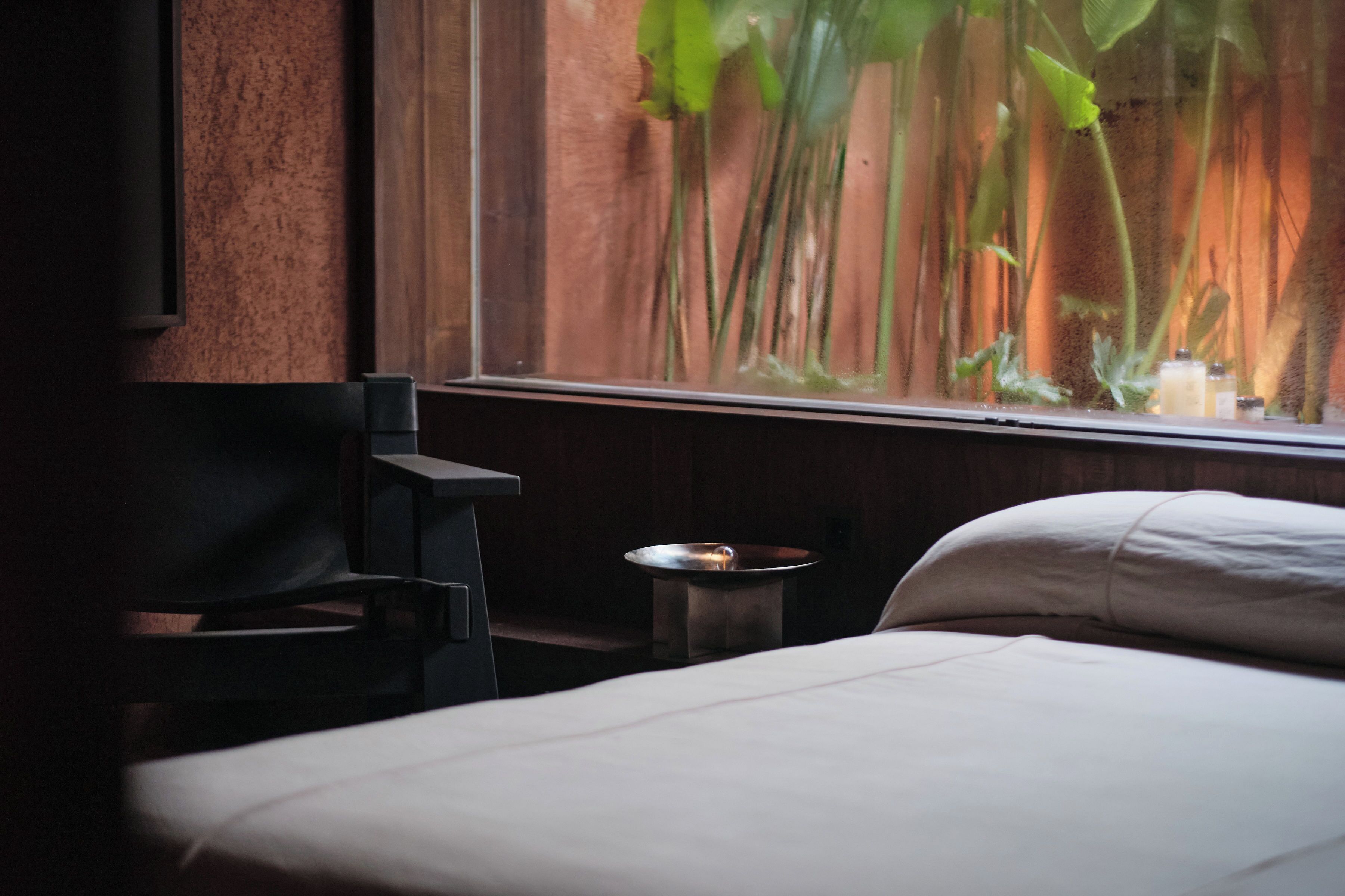 A hotel room with a bed, a small table with a candle, and a window with a view of plants.