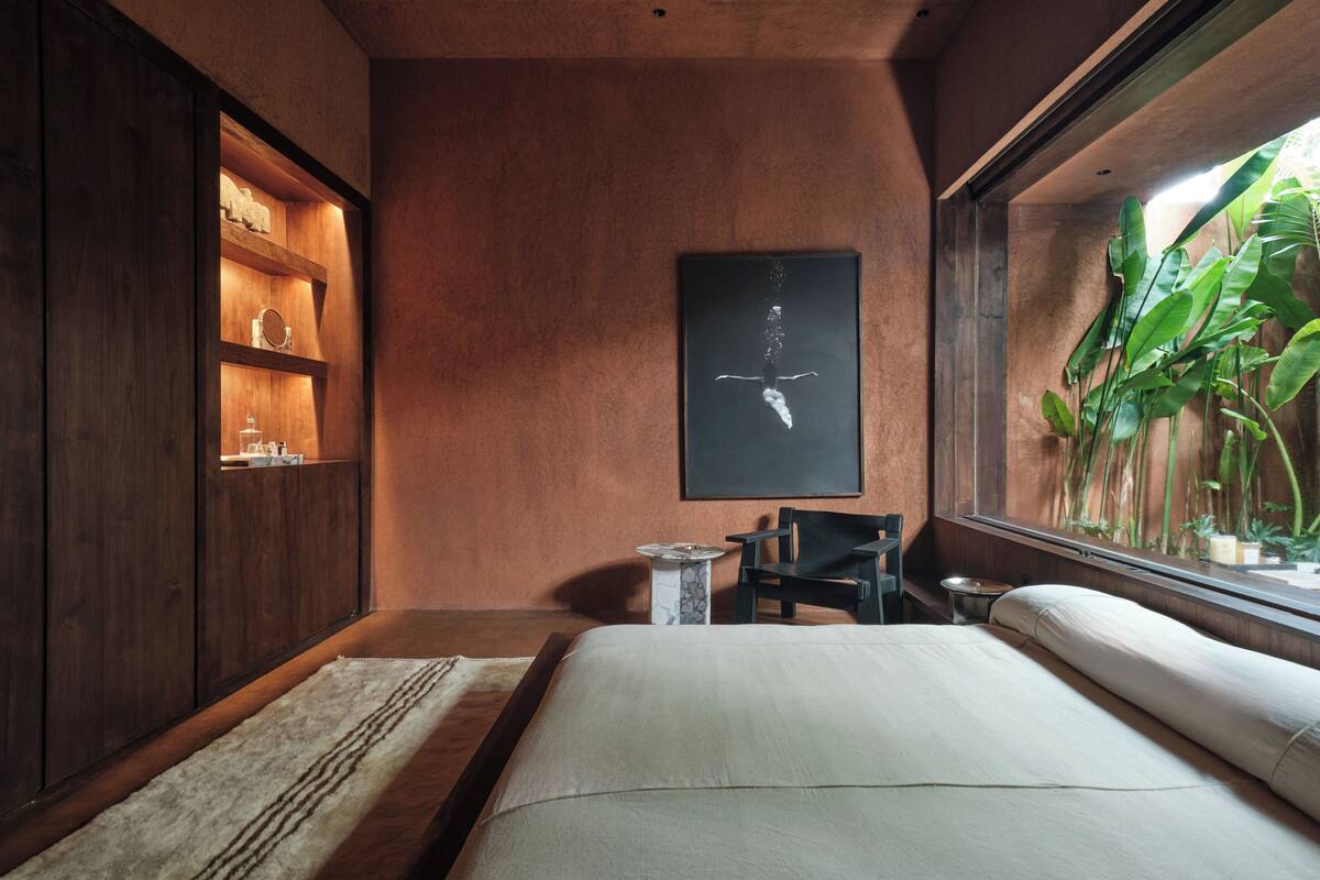 Une chambre à coucher avec un lit, une armoire en bois, une petite table et une fenêtre donnant sur des plantes.