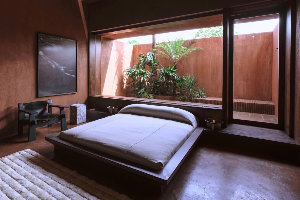 Une chambre à coucher comprenant un lit, une chaise, une petite table, une plante et une fenêtre donnant sur un paysage verdoyant.