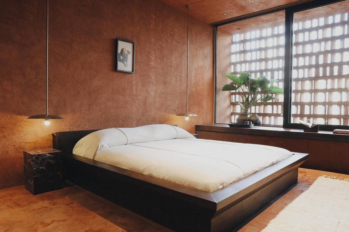 Une chambre moderne avec un grand lit, une fenêtre donnant sur un paysage verdoyant et une petite table de chevet avec une lampe.