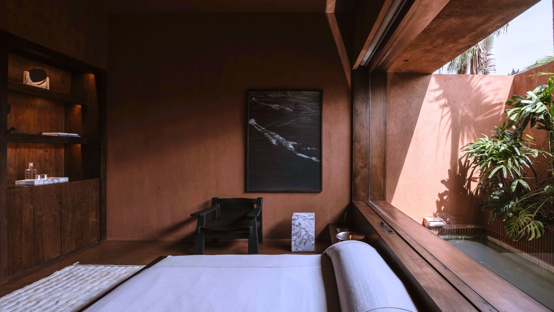 Une pièce comprenant un lit, une chaise, une étagère avec divers objets, un tableau, et donnant sur une piscine et des plantes.