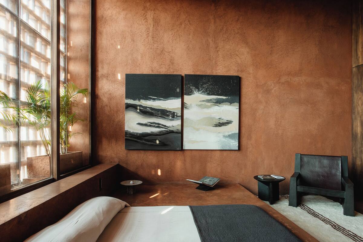 Une chambre à coucher moderne avec un grand lit, une petite table de chevet, une lampe de lecture et un fauteuil. Il y a deux tableaux au mur et une fenêtre avec des plantes.