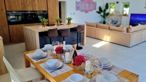 A dining area with a wooden table set for four, surrounded by a modern kitchen and a living room with a sofa and a TV.