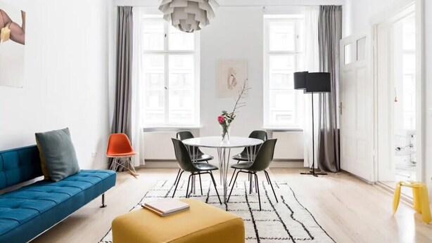 Een moderne woonkamer met een blauwe bank, een geel poufje, een ronde eettafel met stoelen en een opvallende hanglamp.