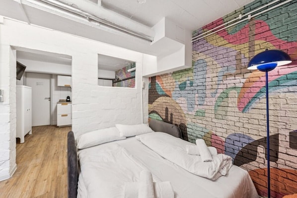 A bedroom with a bed, a white wardrobe, a mirror, a colorful brick wall mural, and a floor lamp.