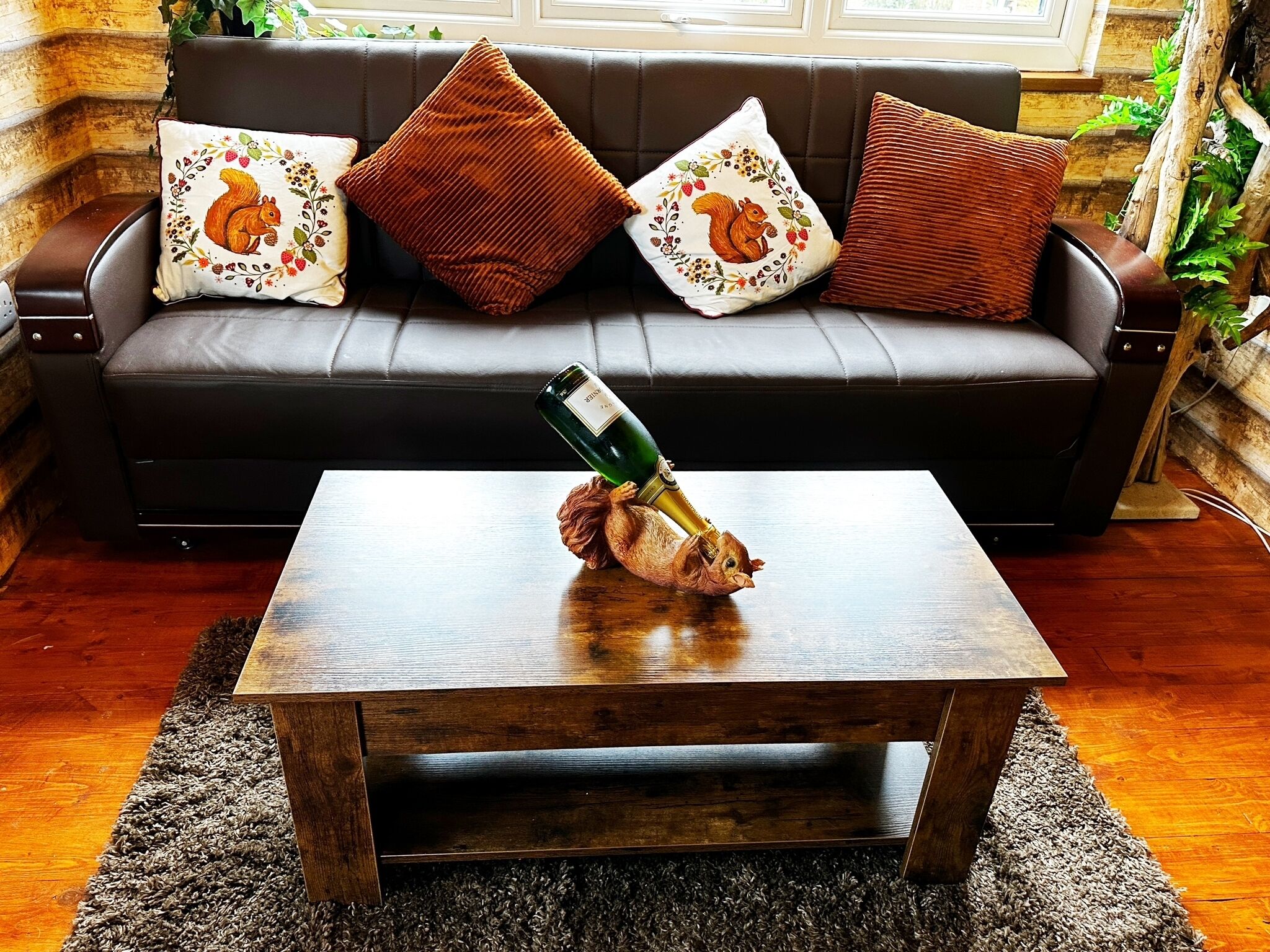 Un salon chaleureux avec une table basse en bois, une bouteille de champagne et un coussin orné d’un motif d’écureuil, posé sur un canapé en cuir foncé.
