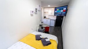 Et lille hotelværelse med seng, skrivebord, tekøkken og et fjernsyn der viser streamingtjenester.