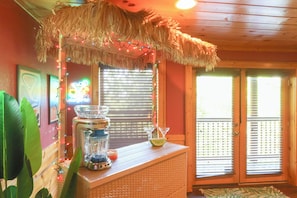 Um quarto com teto de madeira, um telhado de palha sobre um espaço de bar, uma janela com persianas e uma porta com tela treliçada.