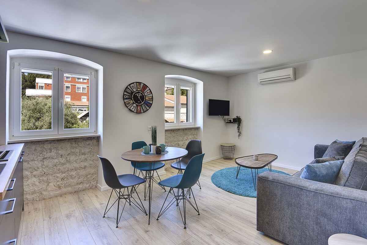 Un salon moderne avec un canapé gris, une table de salle à manger ronde entourée de chaises, un téléviseur fixé au mur et une fenêtre donnant sur des immeubles.