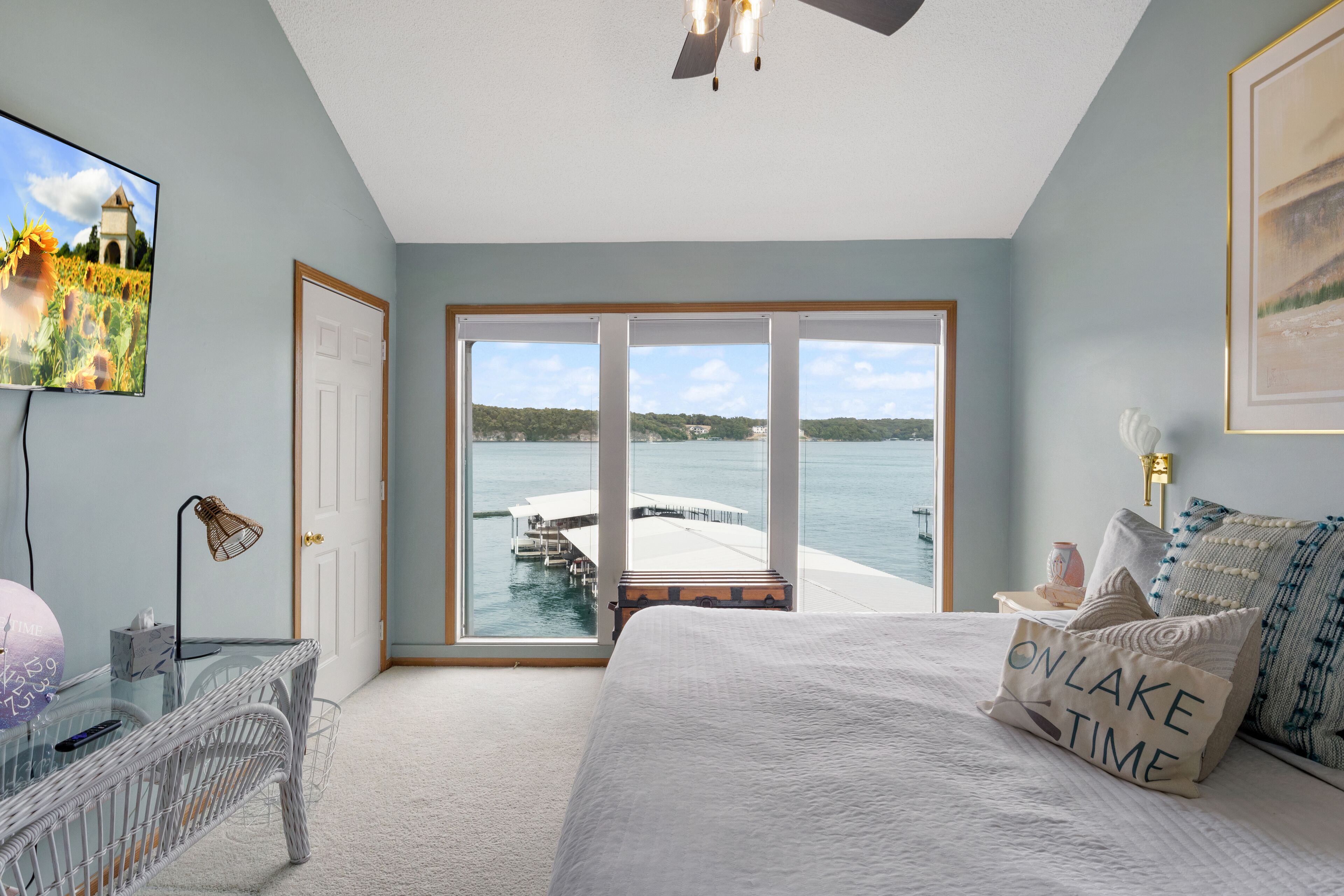 Un dormitorio con una cama grande, una mesa de vidrio, una silla, un televisor y vistas a un lago y un muelle.