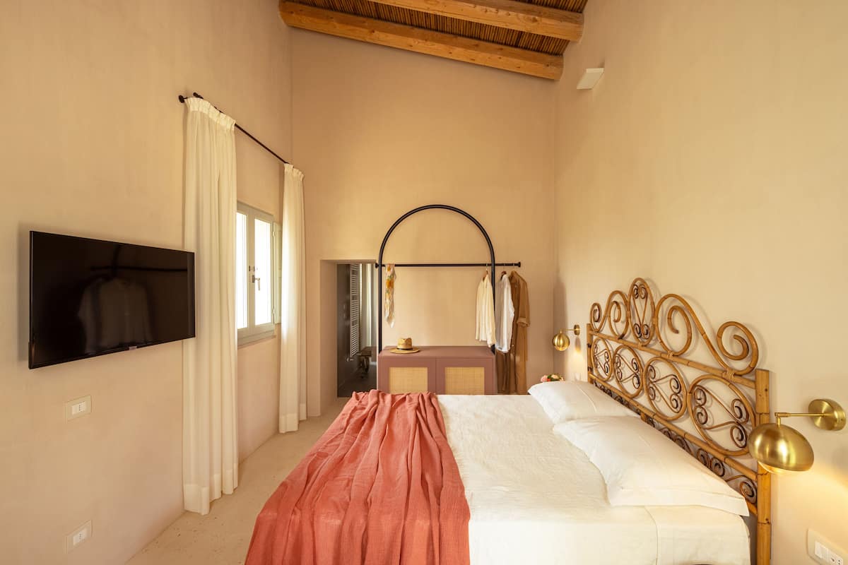 A bedroom with a bed, a TV mounted on the wall, a wooden ceiling, and a window with curtains.