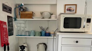 A compact kitchen with a white refrigerator, a microwave, a kettle, and a coffee maker. There are shelves above the fridge holding dishes and a framed picture. - The Beetle Cabin London (Brentford)