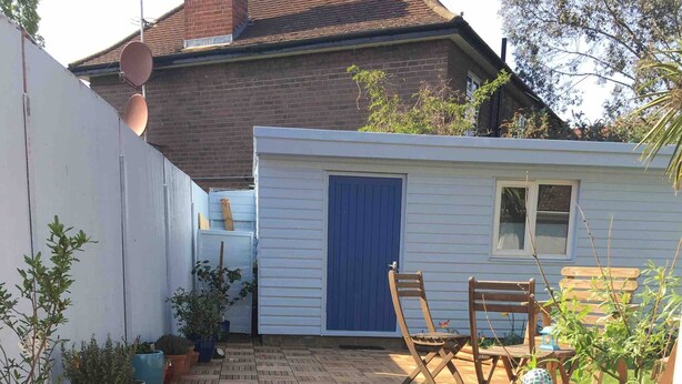 A wooden deck with potted plants, a blue door, and a wooden bench. - The Beetle Cabin London (Brentford)