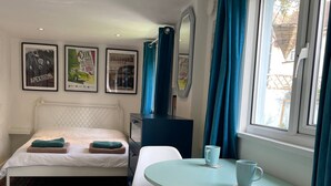 A bedroom with a bed, a desk, two chairs, a window with curtains, and wall art. - The Beetle Cabin London (Brentford)