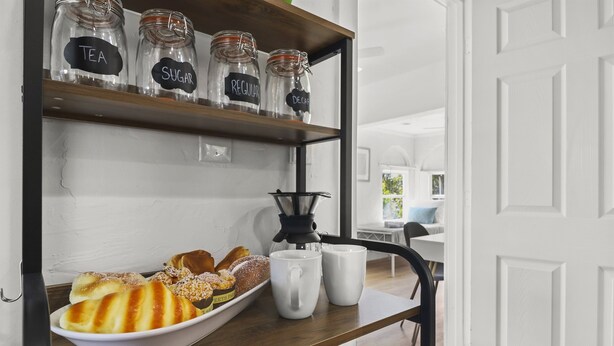 Eine Küchenarbeitsplatte mit Gläsern, beschriftet mit 'Tee', 'Zucker' und 'normal', einer Kaffeemaschine und einer Schüssel Gebäck.