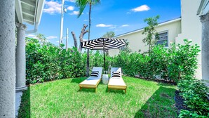 Een verzorgde tuin met twee comfortabele stoelen en een grote parasol, omgeven door weelderige begroeiing en een helderblauwe lucht.