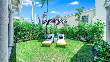 Un jardín bien cuidado con dos sillones de relax y un gran paraguas, rodeado de abundante follaje y un cielo azul despejado.