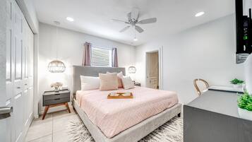 Un dormitorio con cama, mesitas de noche, ventilador de techo, ventana con cortinas y un pequeño escritorio.