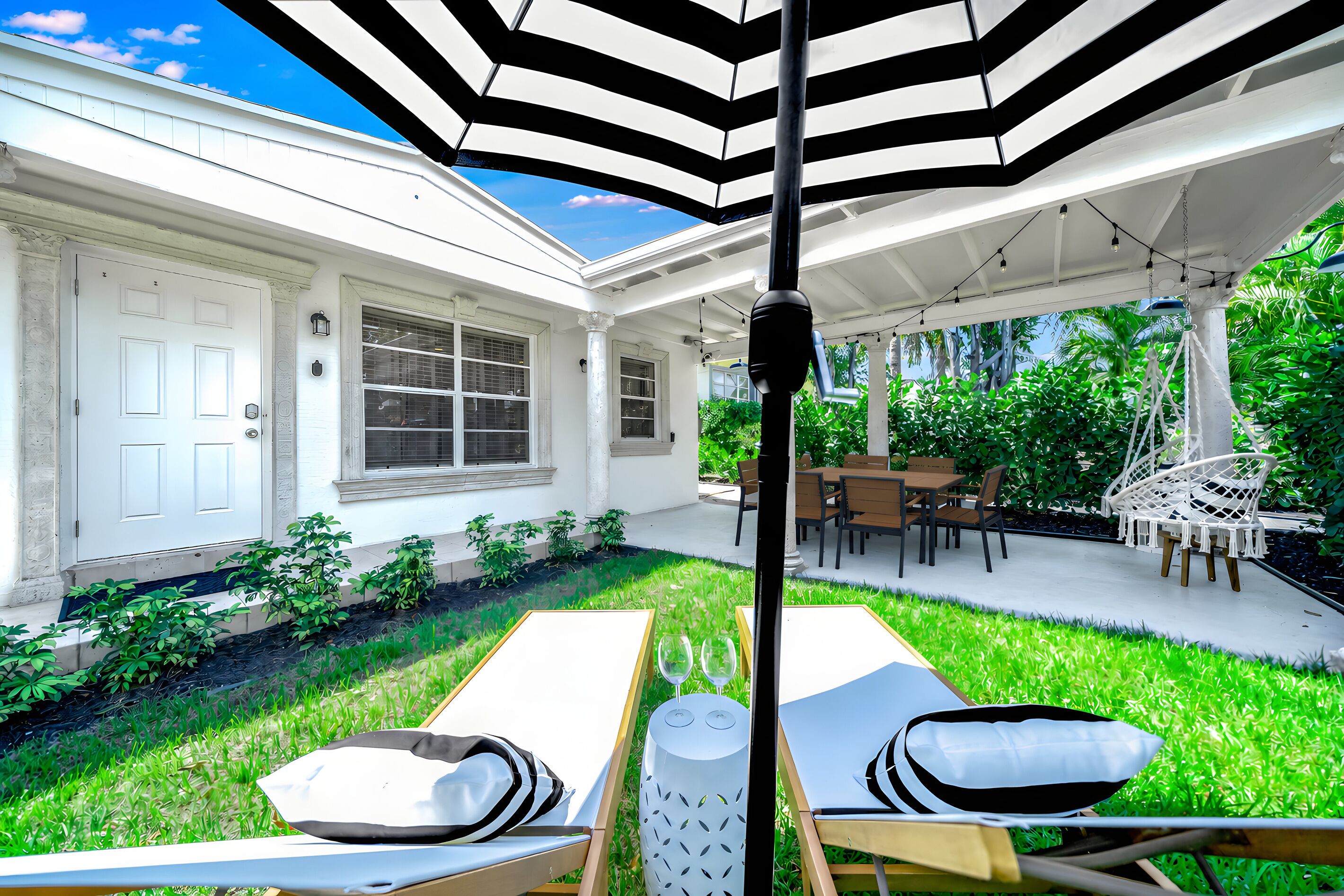A covered patio area with a dining table and chairs, surrounded by lush greenery and a white building with windows.